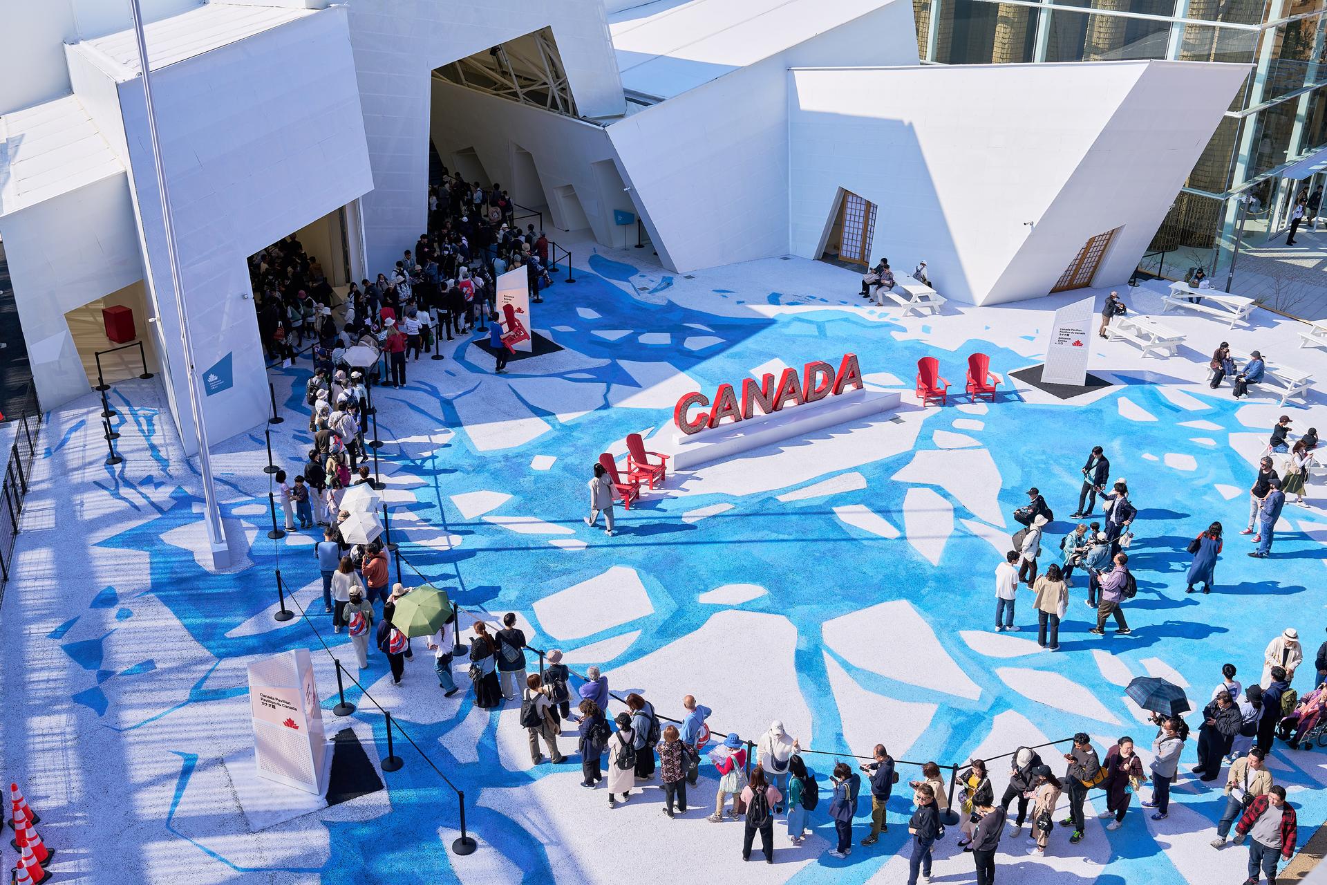 -	An aerial view of the Canada Pavilion at Expo 2025 Osaka reveals a striking white structure resembling a formation of ice. A bold red 'Canada' sign stands prominently at the entrance, while a flowing river design leads the eye toward the pavilion. A long line of visitors eagerly waits to enter the pavilion. Pavillon du Canada à l'Expo 2025 Osaka révèle une structure blanche saisissante ressemblant à une formation de glace. Un panneau rouge « Canada » bien visible se dresse à l'entr