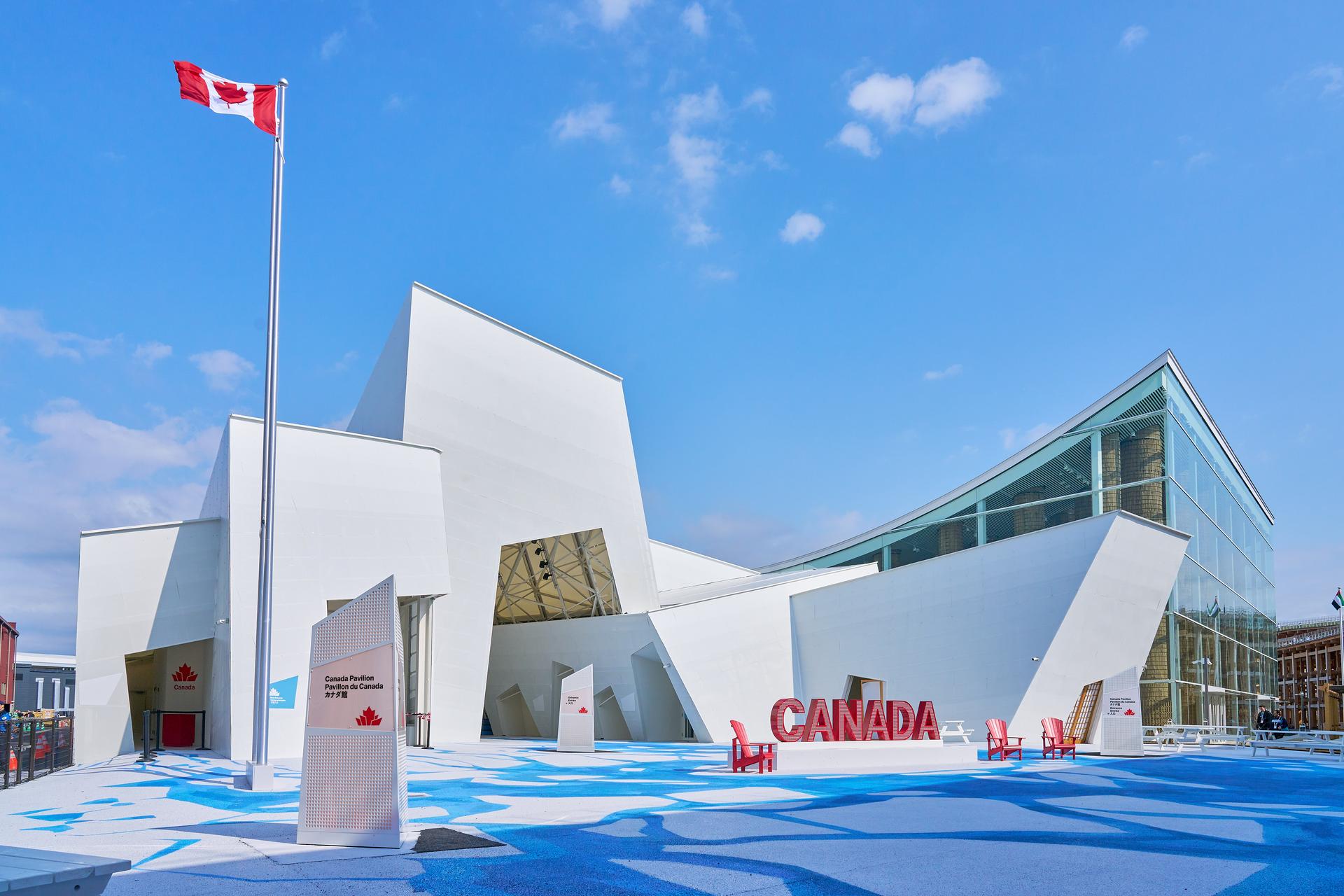 -	An aerial view of the Canada Pavilion at Expo 2025 Osaka reveals a striking white structure resembling a formation of ice. A bold red 'Canada' sign stands prominently at the entrance, while a flowing river design leads the eye toward the pavilion, symbolically drawing visitors in. -	Une vue aérienne du pavillon du Canada à l'Expo 2025 d'Osaka révèle une structure blanche saisissante ressemblant à une formation de glace. Un panneau rouge « Canada » bien visible se dresse à l'entrée.